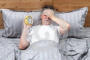 Upset man lying in bed holding yellow alarm clock in hand and making facepalm. Concept when I overslept to work