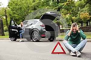 Upset man sitting near warning triangle and broken car