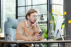 Upset man sitting at the table with a phone in his hands looks tiredly at the computer screen, holds his head with his