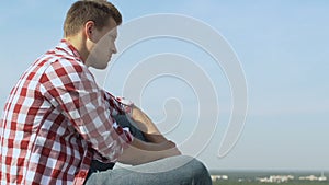 Upset man sitting and yearning on roof of building, looking at city panorama