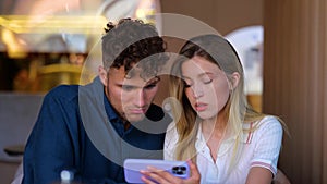 Upset man and woman sitting in cafe and looking at mobile phone screen. Lady holding smart phone near her boyfriend