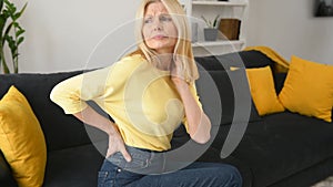 Upset mature 50s woman with blonde-hair suffering from back pain sitting on sofa
