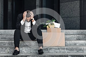 Upset mature businessman sitting with empty poster cardboard sign and box of personal stuff on stairs of office center