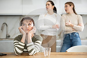 Upset middle-aged woman sitting at the kitchen table with her back to women quarreling to her