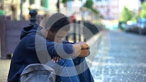 Upset multi-ethnic boy lonely sitting on sidewalk, family conflict, rebellion