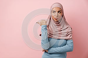 An upset muslim woman wearing pink hijab pointing to the copyspace and wrinkling her forehead