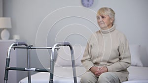 Upset old woman sitting near walking frame, disability after joint surgery