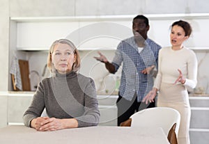 Upset old woman sitting at table with her back to angry middle-aged man and woman