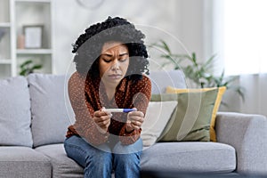 Upset and pensive African American young woman sitting on sofa at home and looking at negative pregnancy test. Ovulation