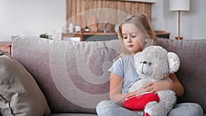 Upset pensive cute little girl hugging bear toy sitting on couch at living room suffering loneliness