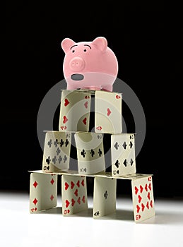 Upset piggy bank standing on fragile shaky house of cards with a dark background