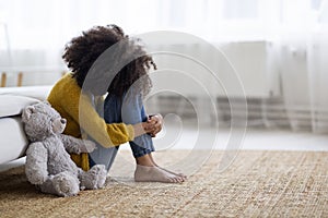 Upset preteen black girl sitting on floor with teddy bear