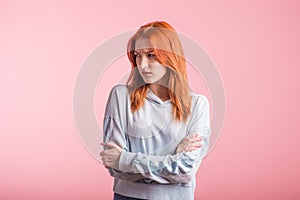 Upset redhead girl with crossed arms in the studio on a pink background.