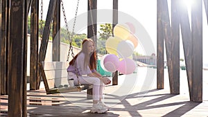 Upset redhead young woman swaying on swing holding hands large fountain of colorful balloons.