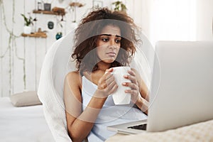 Upset sad beautiful african girl looking at laptop holding cup sitting in chair at home spending her weekend alone.