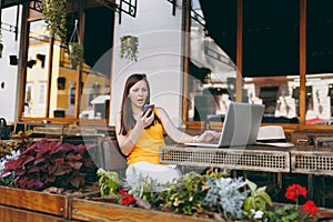 Upset sad girl in outdoors street cafe sitting with laptop pc computer, looking on mobile phone texting message sms