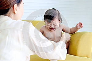 Upset sad toddle baby girl crying on yellow sofa in living room at home, mother from behind try to soothe little kid daughter to photo