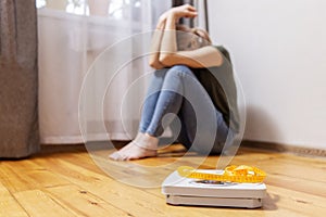 Upset and sad woman sitting on wooden floor near white scale with measuring tape