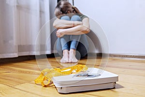 Upset and sad woman sitting on wooden floor near white scale with measuring tape