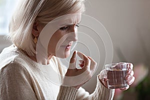 Upset senior woman holding pill and glass water taking medicine