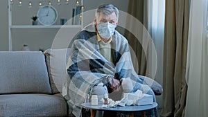 Upset sick grandpa in medical mask sitting on couch caucasian elderly man wrapped in blanket feel chills ill mature