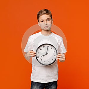 Upset teen boy holding big clock, orange studio background