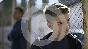 Upset teen boys leaning on metal fence, abandoned with society, orphanage