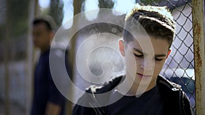 Upset teen boys leaning on metal fence, abandoned with society, orphanage