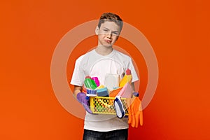 Upset teenage boy standing with lots of detergents and cleaning tools