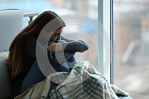 Upset teenage girl sitting at window indoors.