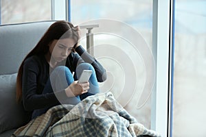 Upset teenage girl with smartphone sitting at window indoors