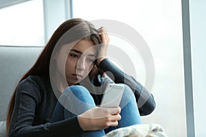 Upset teenage girl with smartphone sitting at window indoors.