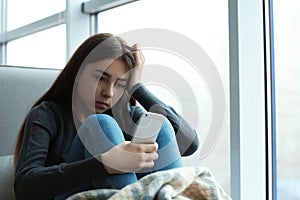 Upset teenage girl with smartphone sitting at window indoors