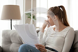 Upset thoughtful woman holding paper document in hands, sitting on sofa