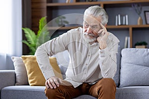 Upset and tired senior gray-haired man sitting on the sofa at home and leaning his head on his hand and looking