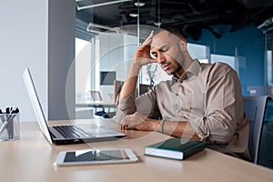 Upset tired young african american man sitting at desk with laptop in office, tired of work holding hand on head