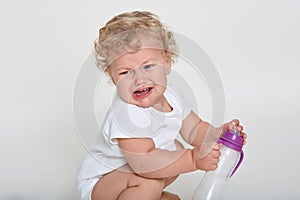 Upset toddler crying while squats against white background, child wants to drink, holds empty sippy cup, infant with beautiful