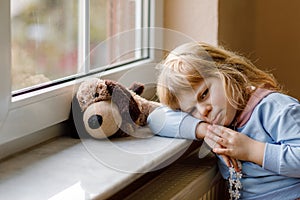 Upset toddler girl sitting by window and looking out. Sad crying child feeling unhappy. Lockdown and isolation concept