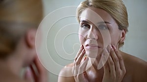 Upset woman looking in mirror and touching face, sad about skin aging, wrinkles