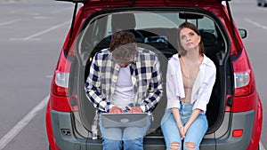 Upset woman sitting on the trunk car next to her boyfriend. Man using laptop while working online. Transportation