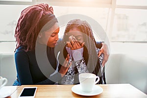 Upset youn african woman sit and cry. She covers face with hands. Her friend comfort young woman. They sit in cafe at photo