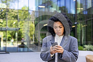 Upset young African American businesswoman standing outside office building and looking worriedly at phone screen. Close