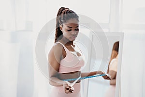 Upset young African American woman measuring her waist near mirror, feeling unhappy with results of weight loss diet