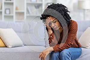 Upset young African American woman sitting on sofa at home, resting head on hand, feeling lonely, suffering from