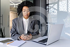 Upset young African American woman working in the office on a laptop, sitting at the desk, holding the phone and looking