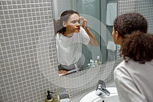 Upset young african woman finding pimple on her face while looking at mirror in bathroom home