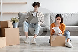 Upset young Asian lady unpacking carton box while her lazy husband sitting on sofa and using laptop, blank space