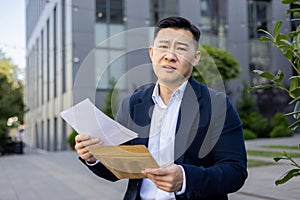 Upset young Asian man in a business suit sitting on a bench outside and looking disappointedly at the camera, holding an