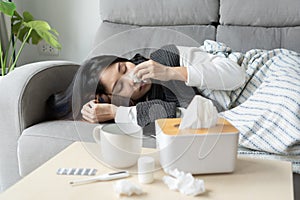 Upset young Asian woman sleeping on couch covered with blanket freezing blowing running nose got fever caught cold sneezing in