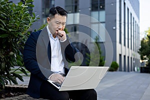 Upset young businessman sitting on a bench near an office center, holding a laptop on his lap and looking worriedly at